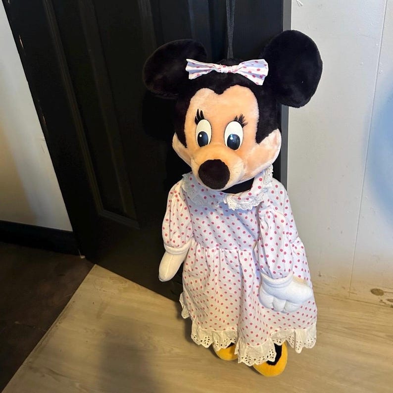 May include: A plush Minnie Mouse doll wearing a pink polka-dot dress with white lace trim and a matching bow. The doll has black ears, a peach-colored face, and yellow shoes. The doll is standing in front of a black door.