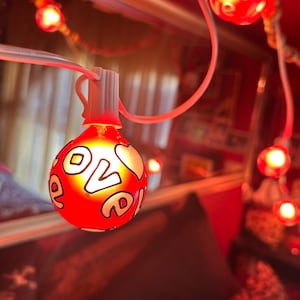 May include: A string of red decorative lights with the word "love" in white script. The lights are shaped like bulbs and have a warm glow. The background is a blurred red.