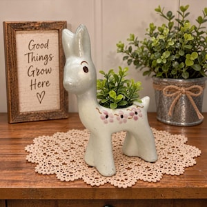 May include: A light green ceramic deer-shaped planter with a speckled finish and painted pink flowers along its back. The deer has upright ears and brown eyes. A small green plant is nestled in the opening on the deer's back.