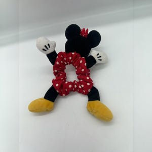 Puede incluir: Un coletero rojo con lunares blancos unido a una figura de peluche. La figura tiene una cabeza negra con orejas redondas, un peque&ntilde;o lazo rojo, guantes blancos y zapatos amarillos. Los brazos y piernas de la figura salen del coletero.