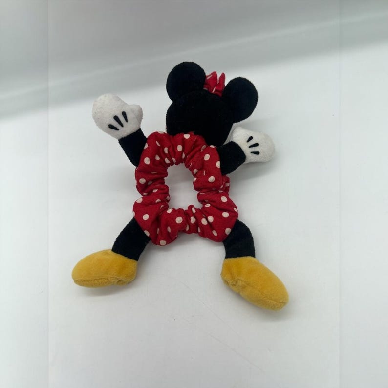 Puede incluir: Un coletero rojo con lunares blancos unido a una figura de peluche. La figura tiene una cabeza negra con orejas redondas, un peque&ntilde;o lazo rojo, guantes blancos y zapatos amarillos. Los brazos y piernas de la figura salen del coletero.