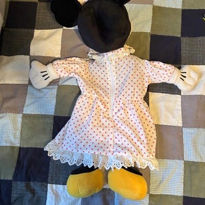 May include: A Minnie Mouse plush doll, featuring black ears, a pink polka dot dress with white lace trim, white gloves with black details, and yellow shoes. The doll is lying on a patchwork quilt.