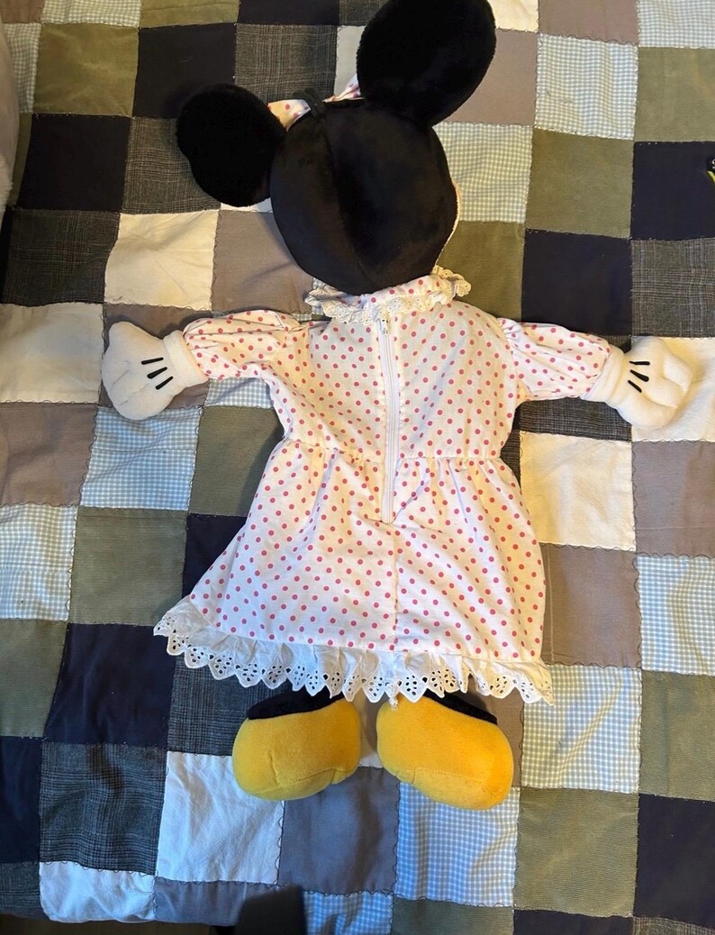 May include: A Minnie Mouse plush doll, featuring black ears, a pink polka dot dress with white lace trim, white gloves with black details, and yellow shoes. The doll is lying on a patchwork quilt.