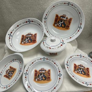 Op de afbeelding: Een verzameling witte keramische serviesstukken met een boerderijscène. Elk stuk heeft een centrale afbeelding van een koe, kip en andere boerderij-elementen in een schuurachtige structuur. De randen zijn versierd met rode harten en blauwe ruitvormen.