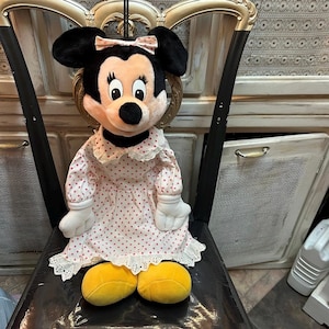 May include: A Minnie Mouse plush doll wearing a pink polka dot dress with white lace trim. The doll has black ears, a pink bow, and yellow shoes. The doll is sitting on a black chair.