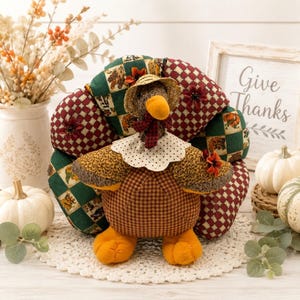 May include: A fabric turkey figurine featuring a round body with brown gingham fabric, a fan-shaped tail made of green and maroon checkered patches, a cream-colored polka dot collar, a small straw hat, and bright orange feet.