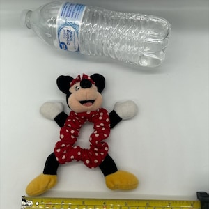 Puede incluir: Un coletero de peluche de Minnie Mouse con una banda de tela roja con lunares blancos, orejas negras, guantes blancos y zapatos amarillos. Una cinta m&eacute;trica amarilla en la parte inferior muestra unos 20 cm de ancho. Una botella de agua arriba.