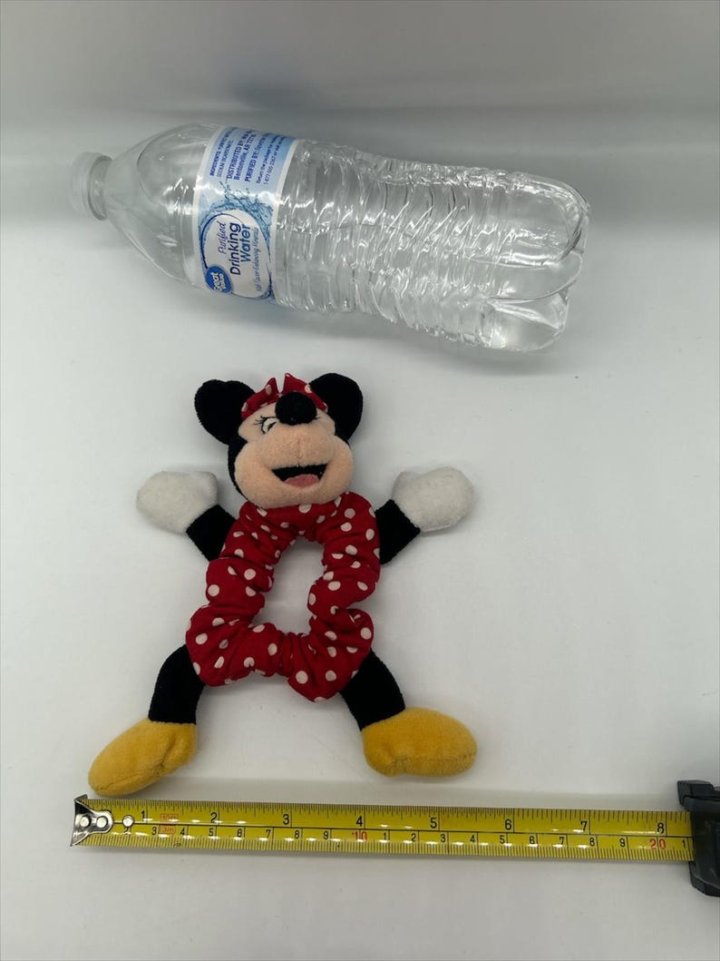 Puede incluir: Un coletero de peluche de Minnie Mouse con una banda de tela roja con lunares blancos, orejas negras, guantes blancos y zapatos amarillos. Una cinta m&eacute;trica amarilla en la parte inferior muestra unos 20 cm de ancho. Una botella de agua arriba.