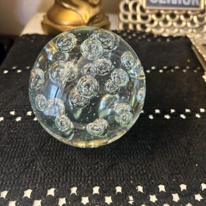 May include: A clear glass paperweight with numerous air bubbles suspended within. The spherical object sits on a black, textured surface with a white, decorative border. The paperweight is a decorative item.