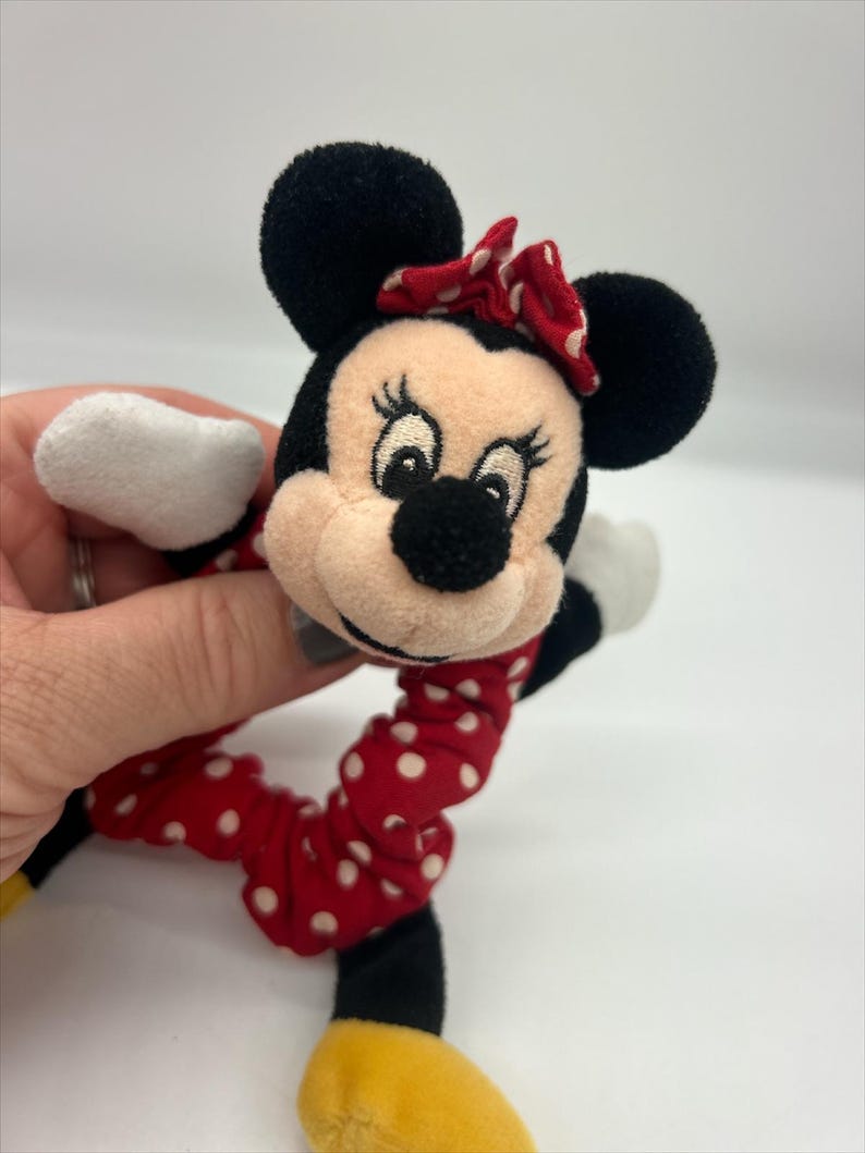 Puede incluir: Un coletero de peluche de Minnie Mouse con una cabeza de tela suave, orejas negras, un lazo rojo con lunares blancos y una banda el&aacute;stica a juego de color rojo con lunares blancos. El personaje tiene ojos, pesta&ntilde;as y nariz negra bordados.