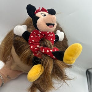 Puede incluir: Un coletero de peluche de Minnie Mouse con tela roja de lunares blancos. El personaje tiene orejas negras, un lazo rojo, guantes blancos y zapatos amarillos unidos a la banda el&aacute;stica, creando un accesorio decorativo para el cabello.