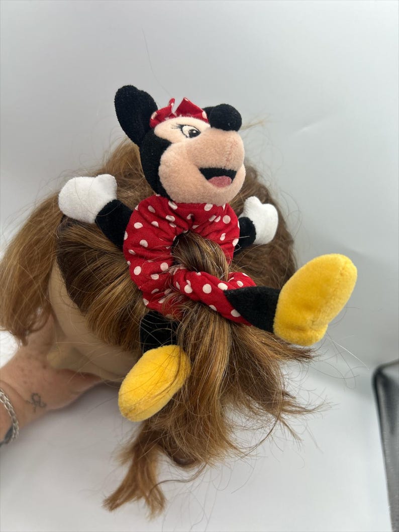 Puede incluir: Un coletero de peluche de Minnie Mouse con tela roja de lunares blancos. El personaje tiene orejas negras, un lazo rojo, guantes blancos y zapatos amarillos unidos a la banda el&aacute;stica, creando un accesorio decorativo para el cabello.