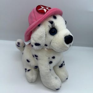 Puede incluir: Un peluche de perro dálmata con manchas negras y ojos azules. El perro lleva un gorro de bombero rosa con un emblema de estrella roja y blanca. El juguete es blanco con manchas negras.