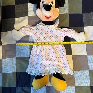 May include: A Minnie Mouse plush doll with a black head, ears, and a pink bow. The doll wears a white dress with pink polka dots and yellow shoes. The doll is lying on a patchwork quilt.