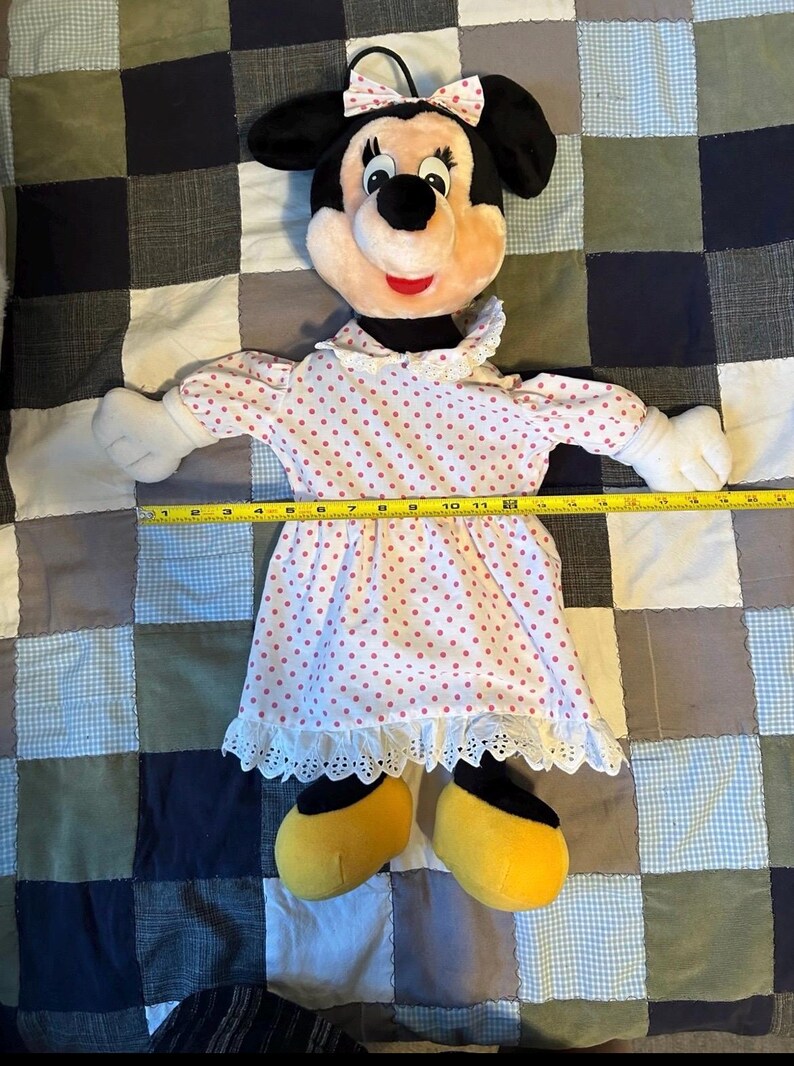 May include: A Minnie Mouse plush doll with a black head, ears, and a pink bow. The doll wears a white dress with pink polka dots and yellow shoes. The doll is lying on a patchwork quilt.