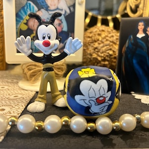 Puede incluir: Una figura del personaje de dibujos animados Wakko Warner, con nariz roja, cuerpo negro, guantes y zapatos blancos y pantalones color caqui. Una pequeña bola redonda azul y amarilla con una cara de personaje de dibujos animados está al lado de la figura. Un collar de perlas con detalles dorados está en primer plano.