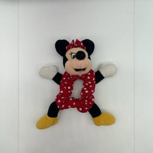 Puede incluir: Un coletero de peluche de Minnie Mouse con una banda de tela roja con lunares blancos. El personaje tiene orejas negras, un lazo rojo con lunares blancos, manos de peluche blancas y pies de peluche amarillos unidos al anillo el&aacute;stico central.
