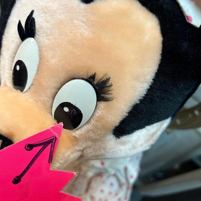 May include: Close-up of a plush toy featuring a character with large, expressive eyes. The toy has a peach-colored face, black hair, and large white eyes with black pupils. A pink tag with a black arrow is in the foreground.