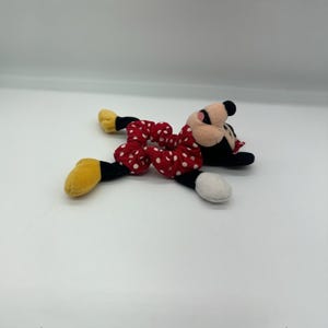 Puede incluir: Un coletero de felpa con tela roja de lunares blancos, unido a una suave cabeza de personaje con orejas negras, cara rosa y pies de felpa amarillos y blancos que se extienden desde los lados de la banda de tela el&aacute;stica.