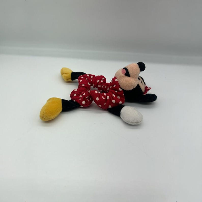 Puede incluir: Un coletero de felpa con tela roja de lunares blancos, unido a una suave cabeza de personaje con orejas negras, cara rosa y pies de felpa amarillos y blancos que se extienden desde los lados de la banda de tela el&aacute;stica.