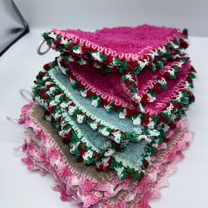 Peut inclure: Une pile de débarbouillettes carrées colorées. Chaque tissu est d'une couleur différente, notamment rose, bleu sarcelle et beige. Les bords sont bordés d'une bordure crochetée rouge, verte et blanche. Chaque tissu a une boucle blanche pour suspendre.