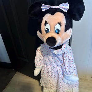 May include: A plush Minnie Mouse doll with black ears, a pink face, and blue eyes. She wears a pink dress with red polka dots, a white lace collar, and a matching bow. The doll has white gloves and yellow shoes.