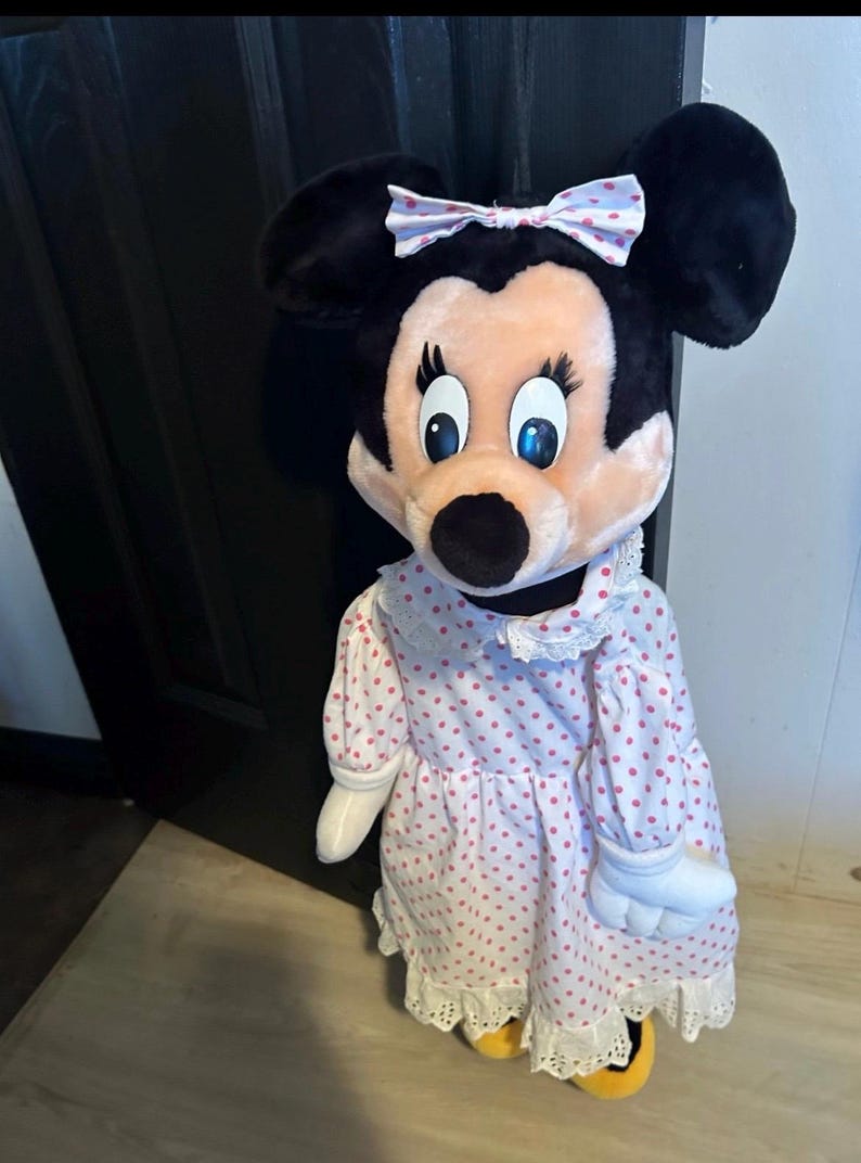 May include: A plush Minnie Mouse doll with black ears, a pink face, and blue eyes. She wears a pink dress with red polka dots, a white lace collar, and a matching bow. The doll has white gloves and yellow shoes.