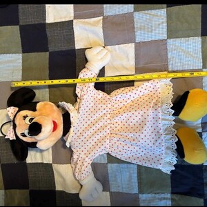 May include: A Minnie Mouse plush doll, approximately 24 inches long, lies on a patchwork quilt. The doll features a pink and white polka-dot dress with lace trim, white gloves, and yellow shoes. Minnie's iconic black ears and bow are visible.