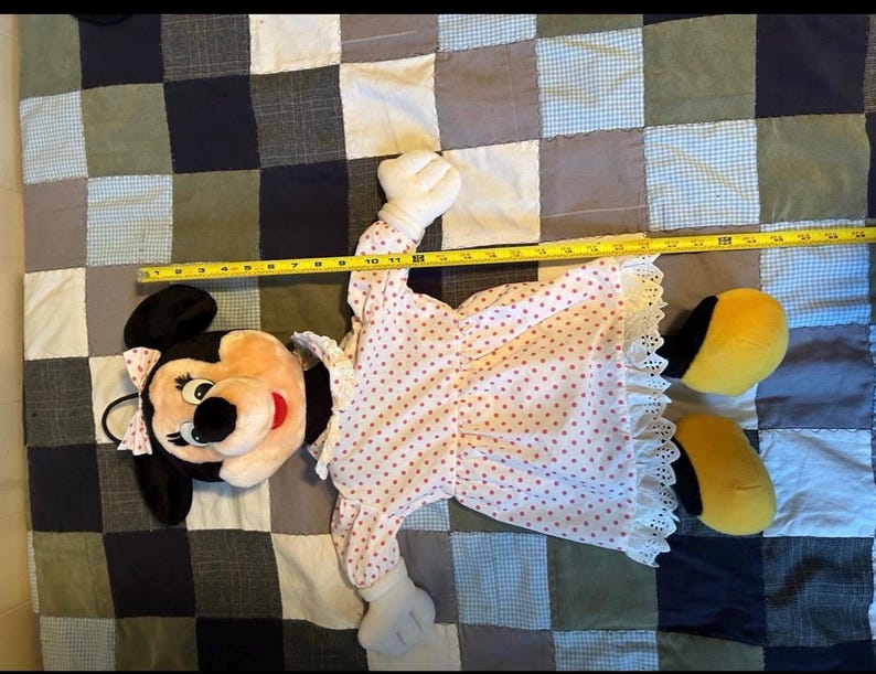 May include: A Minnie Mouse plush doll, approximately 24 inches long, lies on a patchwork quilt. The doll features a pink and white polka-dot dress with lace trim, white gloves, and yellow shoes. Minnie's iconic black ears and bow are visible.