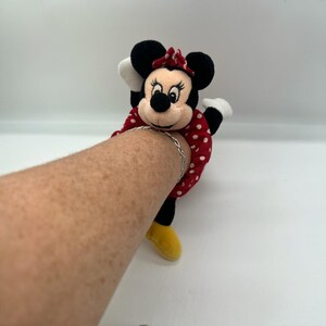 Puede incluir: Un juguete de peluche de Minnie Mouse con un vestido rojo de lunares blancos, un lazo rojo a juego, orejas negras y zapatos amarillos. El juguete est&aacute; dise&ntilde;ado para envolverse alrededor de un brazo, con una pulsera de cadena de metal plateado visible.