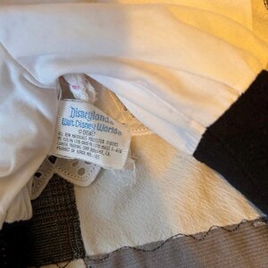 May include: Close-up of a white fabric with a Disneyland and Walt Disney World tag. The tag is rectangular with blue text and a white background. The fabric is layered with a black sleeve visible.