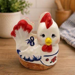 May include: A white ceramic chicken-shaped planter or holder with a red comb, yellow beak, and blue painted details. The base features a brown textured basket-weave pattern, while the side displays a blue bow and a small red floral accent.