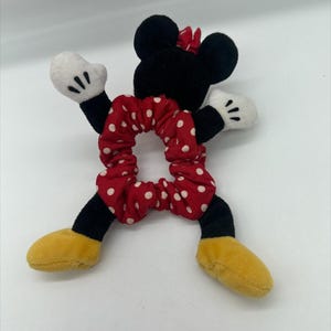 Puede incluir: Un coletero de felpa con tela roja de lunares blancos, unido a una figura suave con orejas negras, guantes blancos y zapatos amarillos. El personaje tiene un lazo rojo sobre la cabeza y brazos y piernas de color negro.
