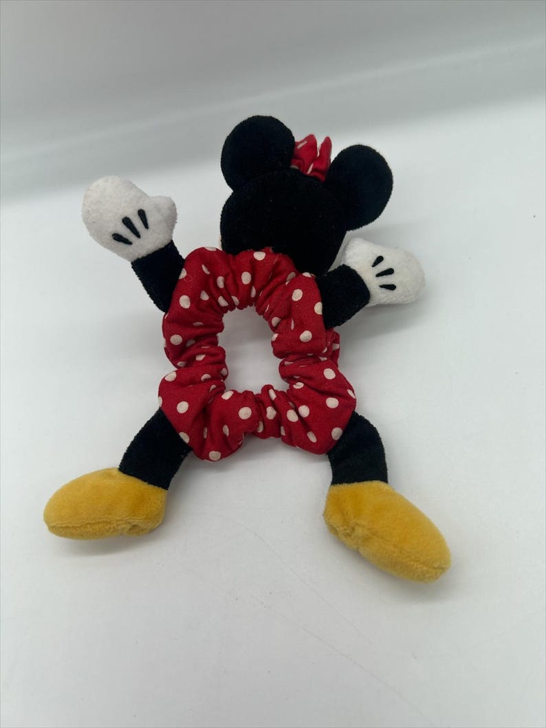 Puede incluir: Un coletero de felpa con tela roja de lunares blancos, unido a una figura suave con orejas negras, guantes blancos y zapatos amarillos. El personaje tiene un lazo rojo sobre la cabeza y brazos y piernas de color negro.