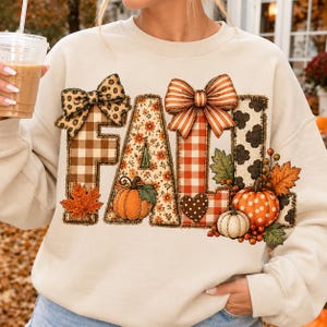 May include: A cream-coloured crewneck sweatshirt featuring a large graphic that spells out the word FALL. The letters are decorated with various patterns including leopard print, brown plaid, floral, orange gingham, and cow print, accented with pumpkins.