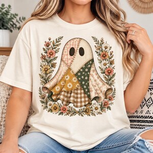 Peut inclure: Un t-shirt couleur crème arborant un motif central représentant un fantôme composé de divers tissus en patchwork, incluant des imprimés à carreaux, floraux et vichy dans des tons terreux, entouré d'une couronne de marguerites et de fleurs.
