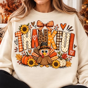 Può includere: Una felpa color crema con una stampa grafica della parola THANKFUL in vari motivi, tra cui scacchi, stampa leopardata e pois. Un tacchino dei cartoni animati con un cappello da pellegrino, girasoli e zucche circondano il testo.