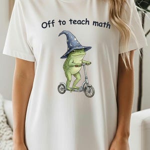 May include: White t-shirt with the text "Off to teach math." The shirt features a whimsical illustration of a green frog wearing a wizard hat and riding a scooter. The frog is the central focus of the design.