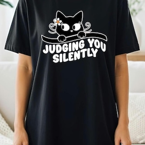 May include: Black t-shirt featuring a cartoon cat with a flower and the text "JUDGING YOU SILENTLY" in white. The cat has large eyes and is peeking over a curved line. The shirt is a casual, comfortable style.