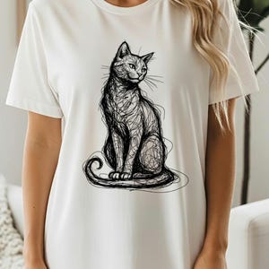 May include: White t-shirt featuring a black sketch of a cat. The cat is depicted in a sitting position with its tail curled. The design is a line drawing style, giving it a simple yet artistic look. The shirt has short sleeves and a crew neck.