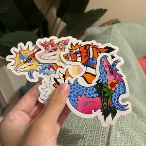 May include: A collection of four die-cut vinyl stickers featuring stylized anthropomorphic animal characters. The designs include vibrant colors, floral accents, sun motifs, and intricate patterns, each with a distinct color palette and character expression.
