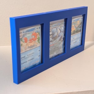 Op de afbeelding: Een blauw frame met drie ruilkaarten. De kaarten tonen kleurrijke illustraties van verschillende personages. Het frame is effen, levendig blauw en de kaarten zijn zichtbaar door rechthoekige openingen.