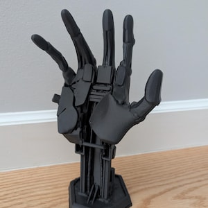Könnte beinhalten: Eine schwarze, bewegliche Handskulptur, 3D-gedruckt mit komplizierten Details. Die Hand ist mit ausgestreckten Fingern posiert und auf einem geometrischen Sockel montiert. Die Skulptur wird auf einer Holzoberfläche vor einer weißen Wand ausgestellt.