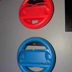 Könnte beinhalten: Zwei Lenkrad-Zubehörteile aus Kunststoff, eines rot und eines blau, für eine Spielkonsole. Jedes Rad hat eine zentrale Halterung für einen Controller und ein schwarzes Akzentstück. Sie sind an einer grauen Wand befestigt.
