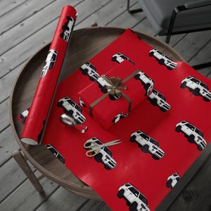 May include: Red wrapping paper with a repeating pattern of white and black cars. A wrapped gift box with a brown ribbon, scissors, and a roll of matching paper are on a wooden table.