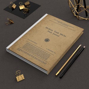 May include: A vintage-style journal with the text "Medium Tank M4A6 Daily Journal" on the cover. The cover has a distressed, aged appearance. Gold binder clips and pencils are arranged around the journal on a dark surface.