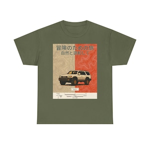 May include: Olive green t-shirt featuring a graphic of a white SUV with a red and tan background. The graphic includes Japanese text and a topographic map design. The shirt is made of a soft, comfortable material.