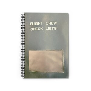 May include: A spiral-bound notebook with the text "FLIGHT CREW CHECK LISTS" in white. The cover is a muted gray-green with a clear pocket. The notebook has a black spiral binding.