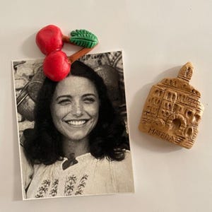 Peut inclure: Une photo d'une femme souriante avec un aimant cerise rouge et un aimant Matera marron. L'aimant cerise a deux cerises rouges et une feuille verte. L'aimant Matera est marron et porte l'inscription "MATERA".