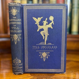 May include: A blue hardcover book titled The Marvellous Adventures of Tyll Owlglass with gold embossed lettering and a central illustration of a jester holding an owl and a mirror. The spine features decorative gold filigree and text details.